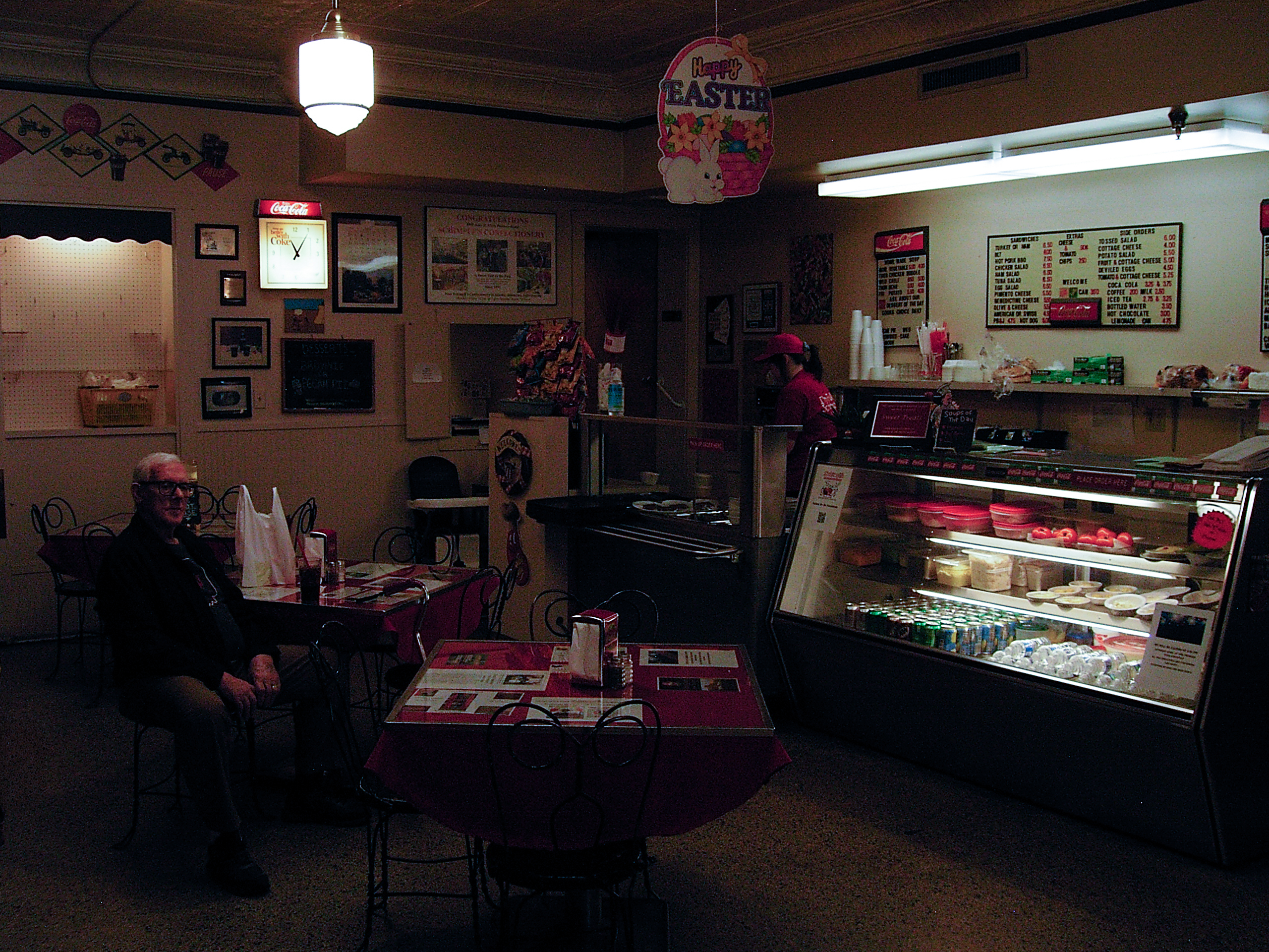 A dim diner interior — a man waits at a small table near the pastry case beneath an Easter decoration