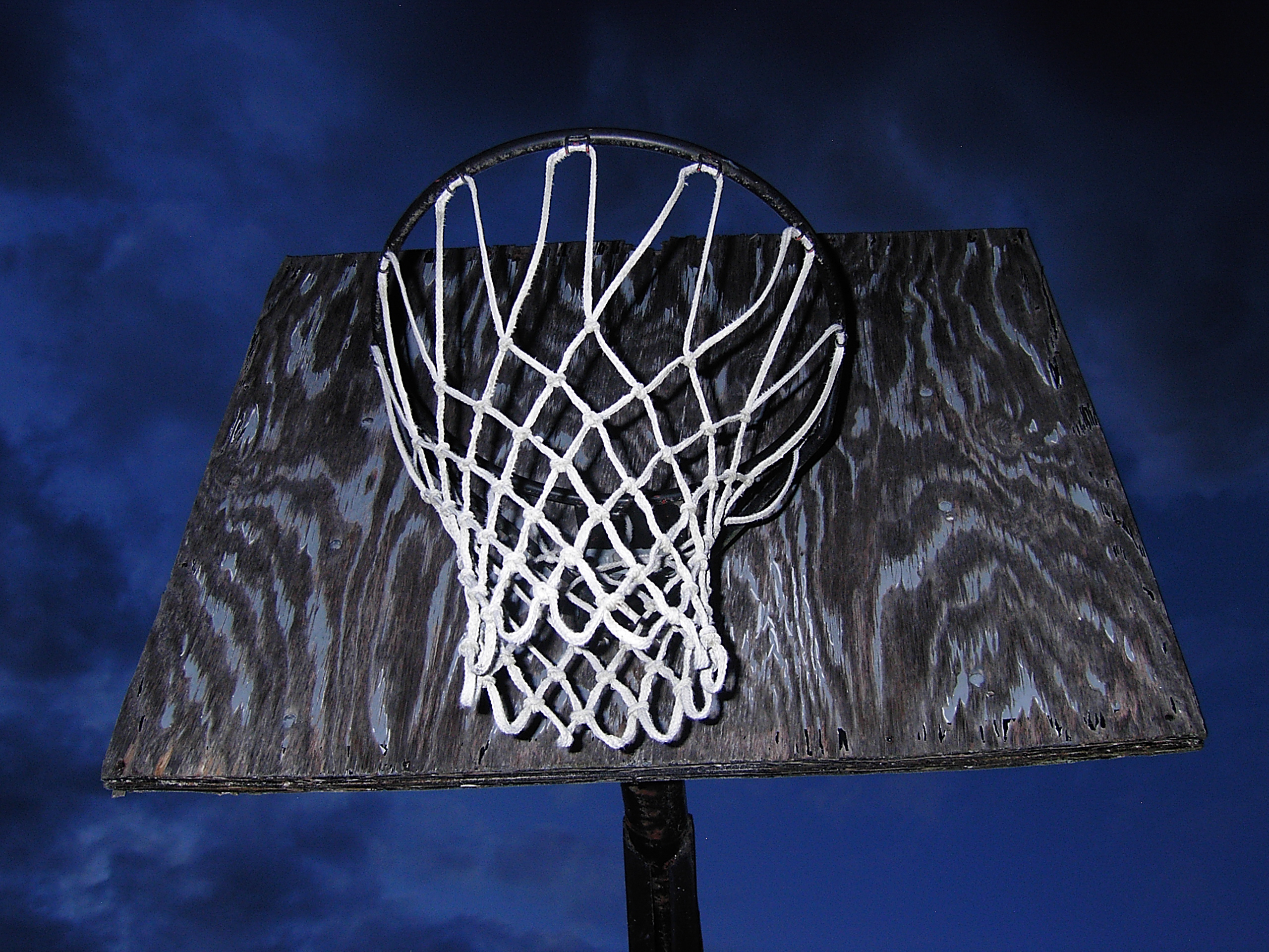 A netted hoop on a weathered plywood backboard, seen from below against a darkening sky