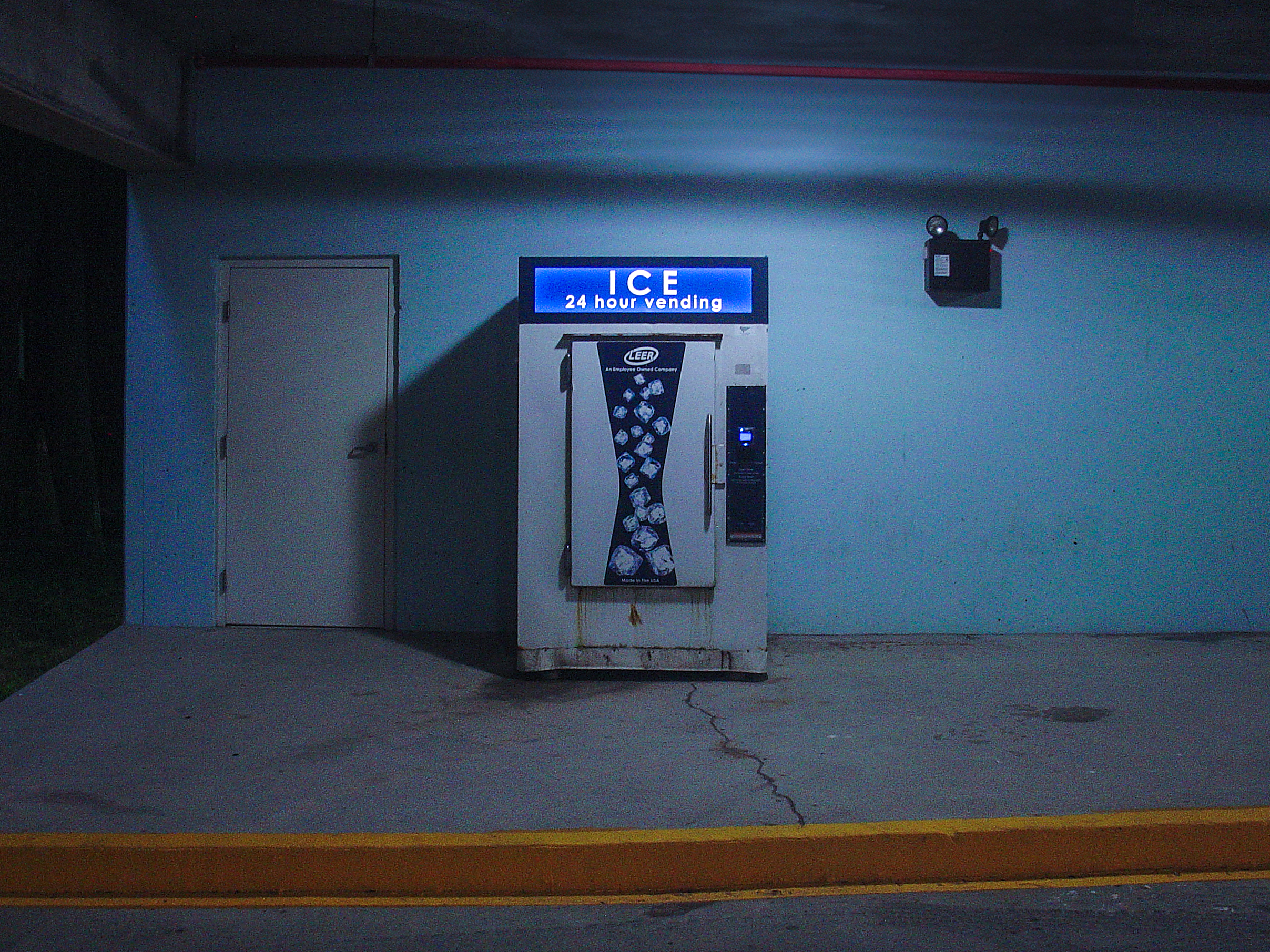 A glowing ice vending machine against a teal wall at night, a white door to its left, a yellow curb crossing the foreground