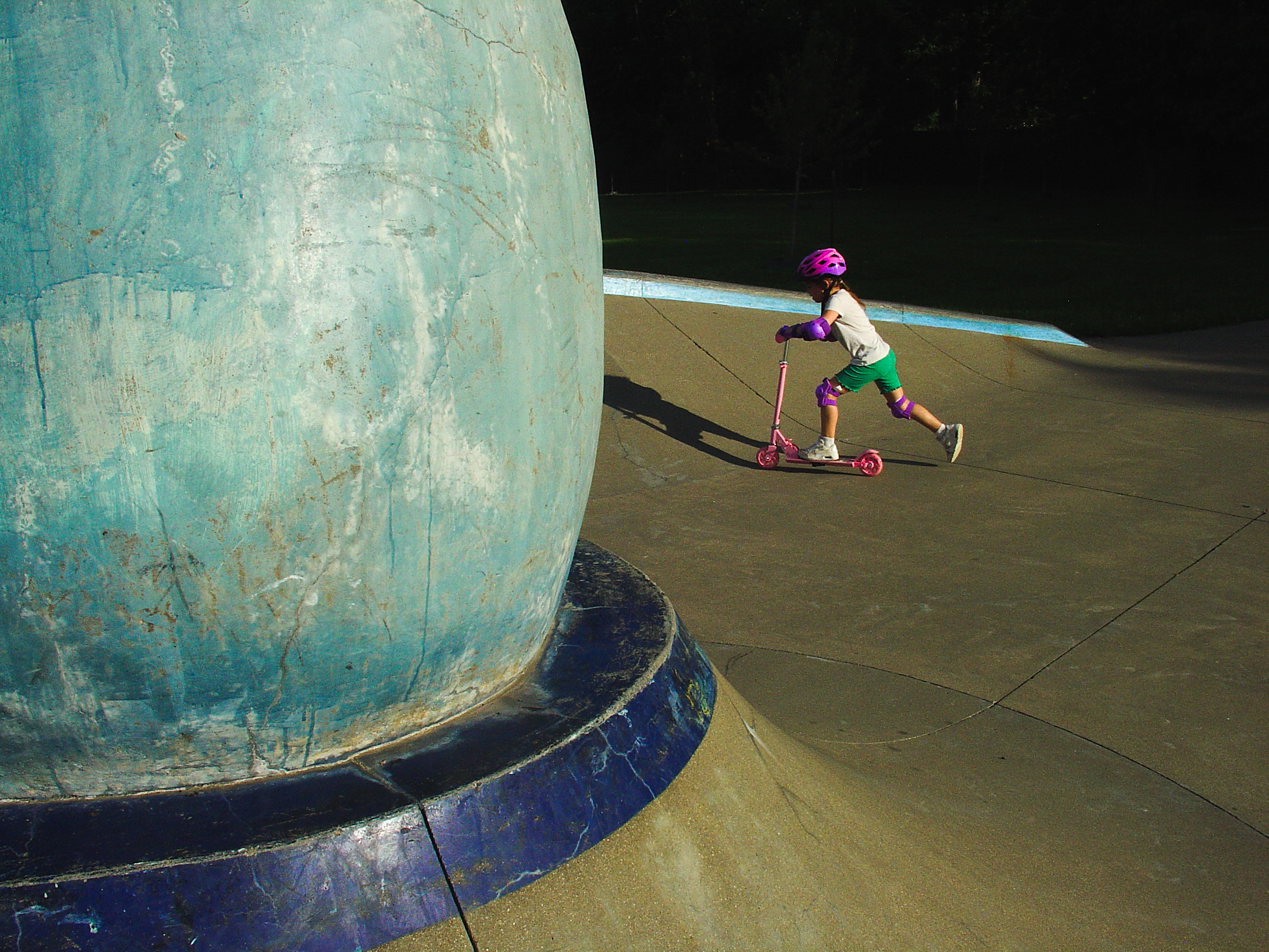 A child on a scooter crossing a sunlit splash pad, the weathered turquoise base of a fountain filling the left of the frame