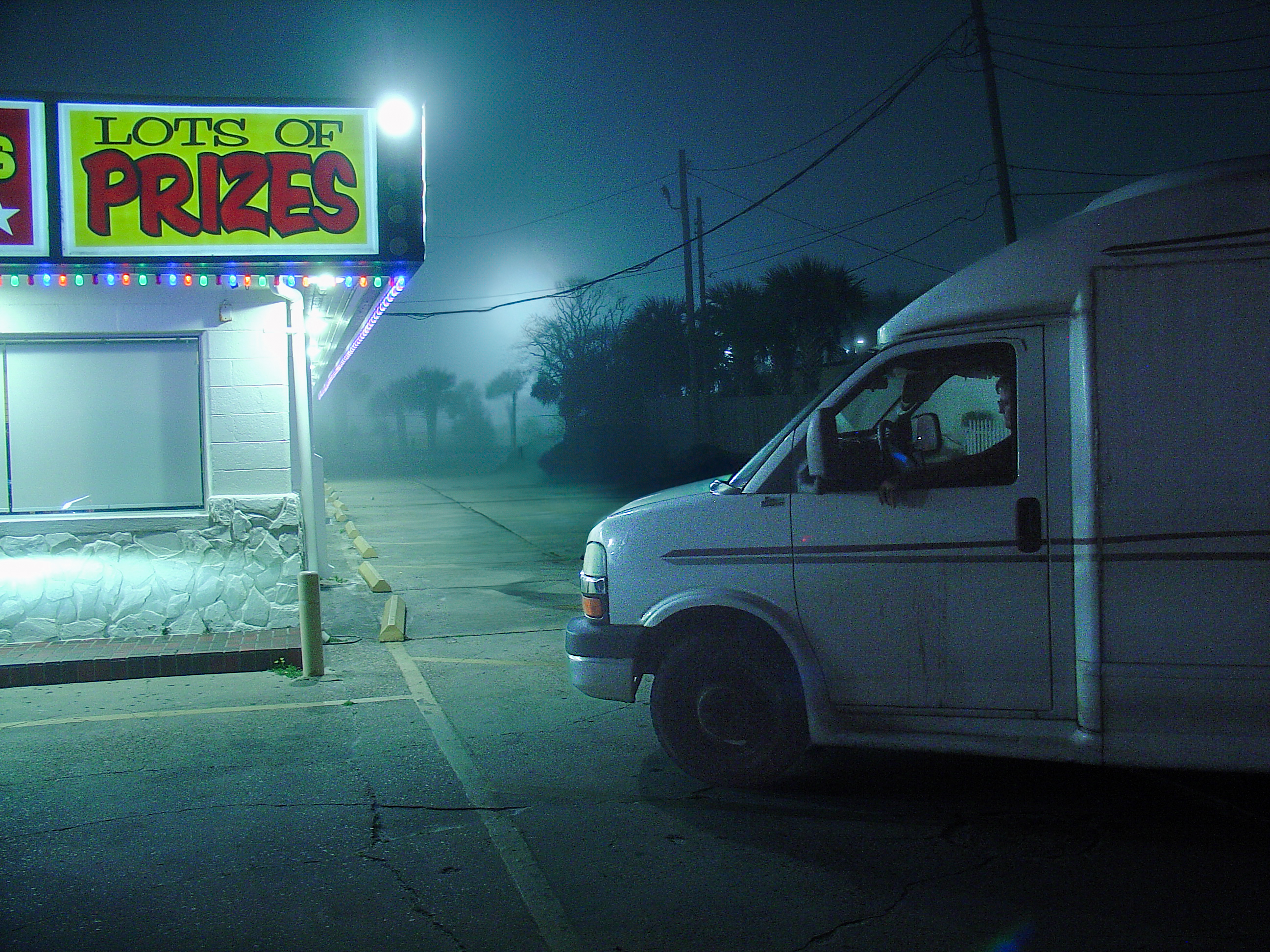 A glowing lots-of-prizes sign over a white van idling in fog, a figure at the wheel, empty lot behind