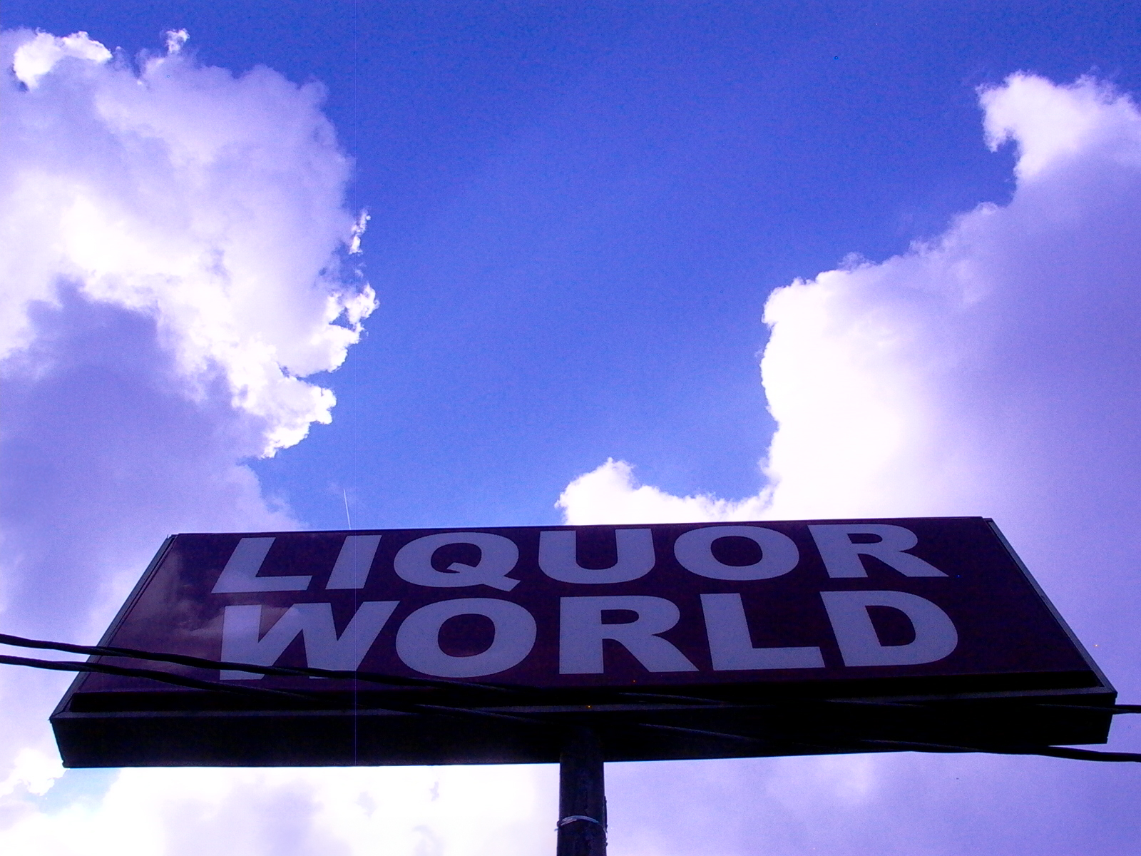A liquor-world sign shot from below against towering white clouds and deep blue sky