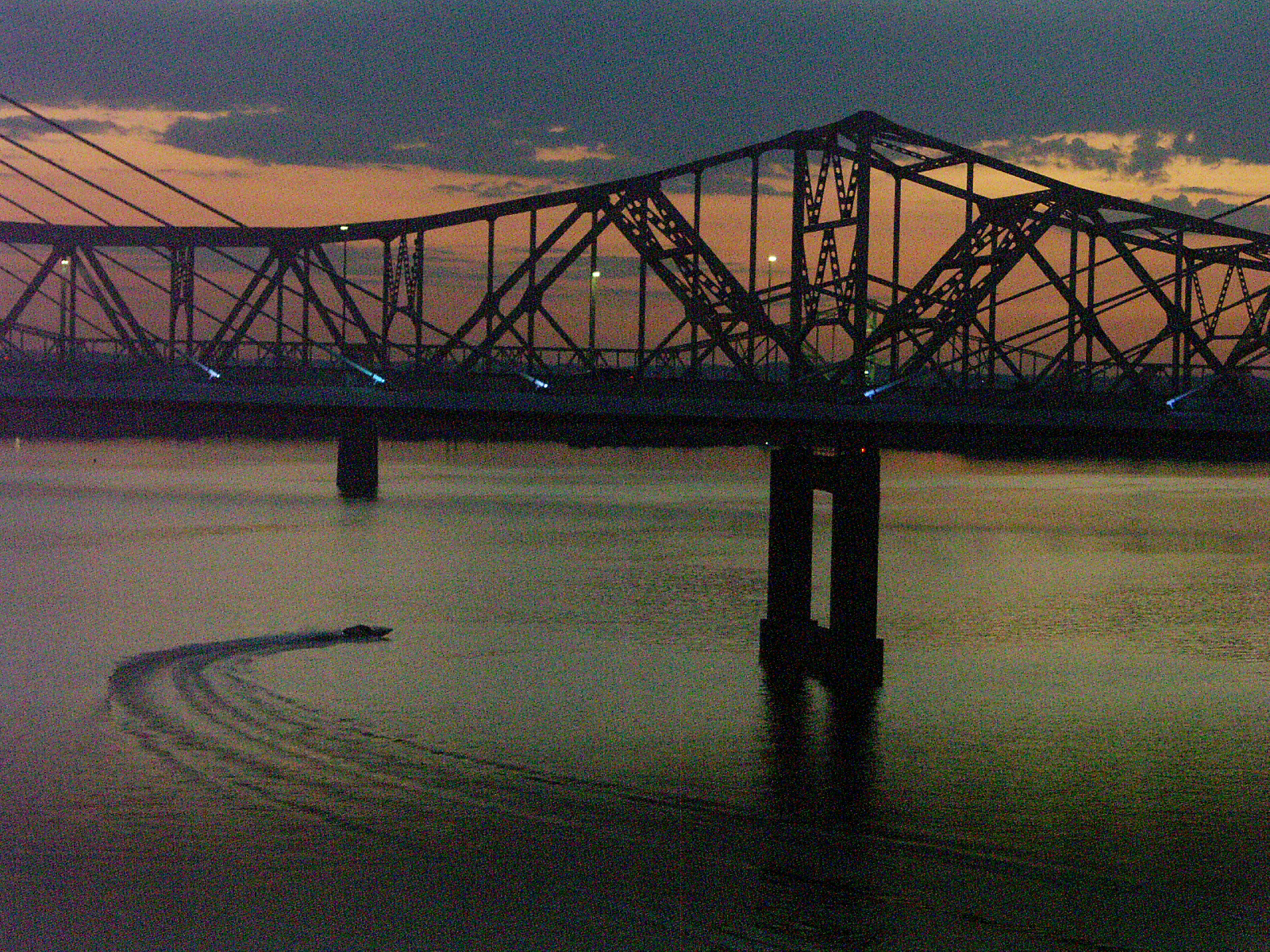 A steel truss bridge silhouetted at dusk over a river, a small boat drawing a curved wake across the water