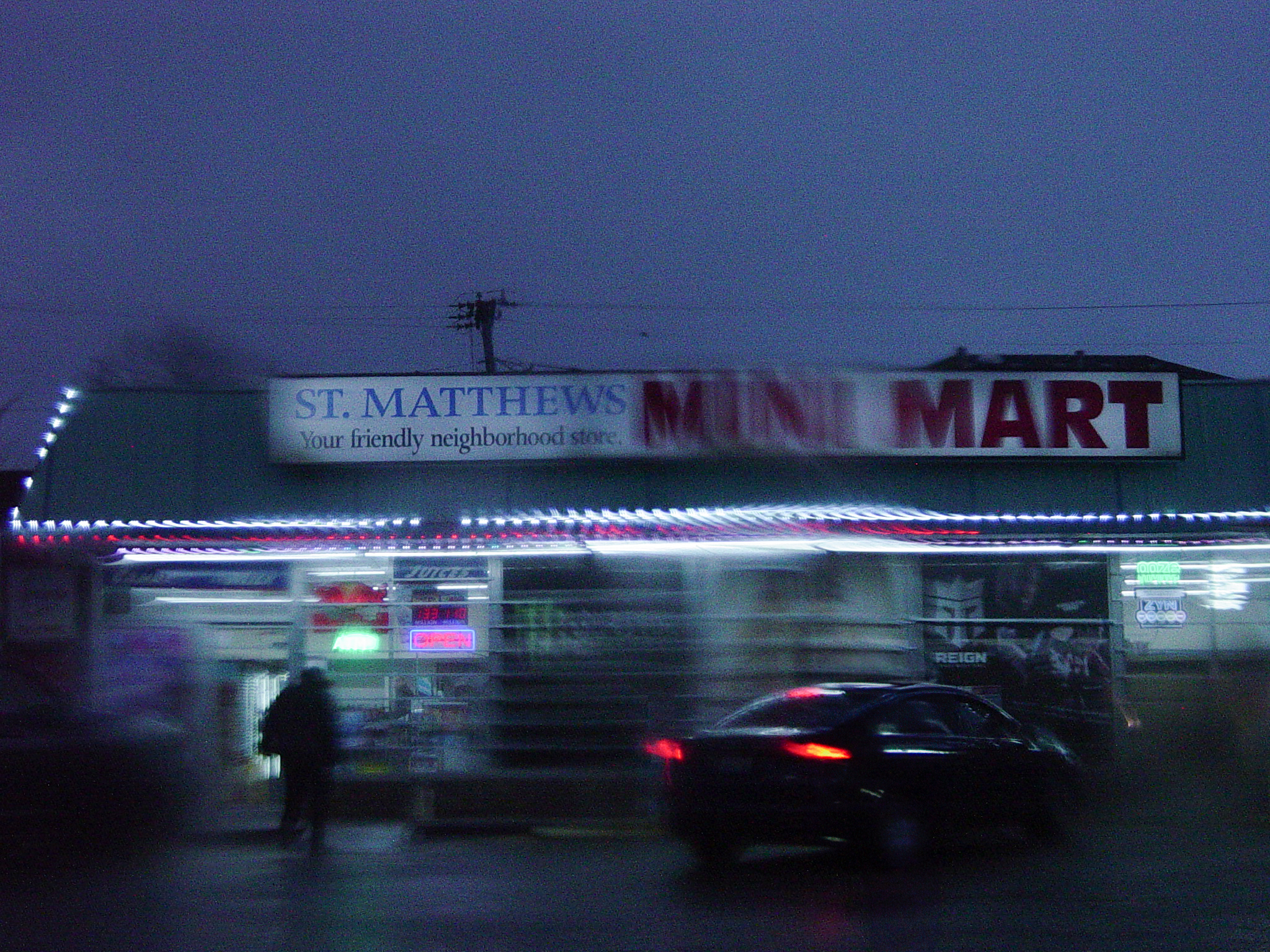 A mini mart at dusk, car lights streaking in front of the blue storefront and red sign