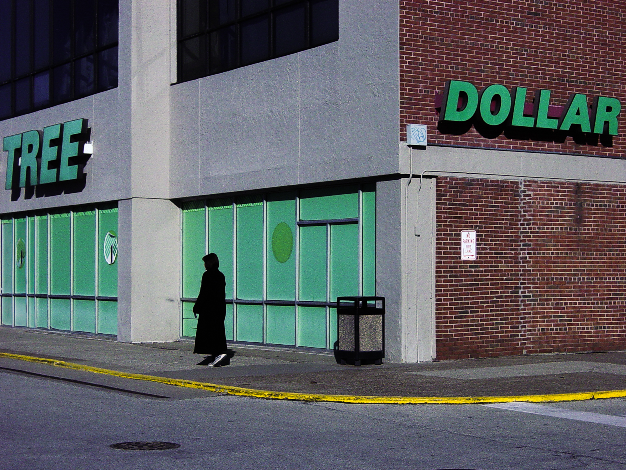 A small figure walking past a Dollar Tree wall, the green storefront panels glowing against brick and pavement