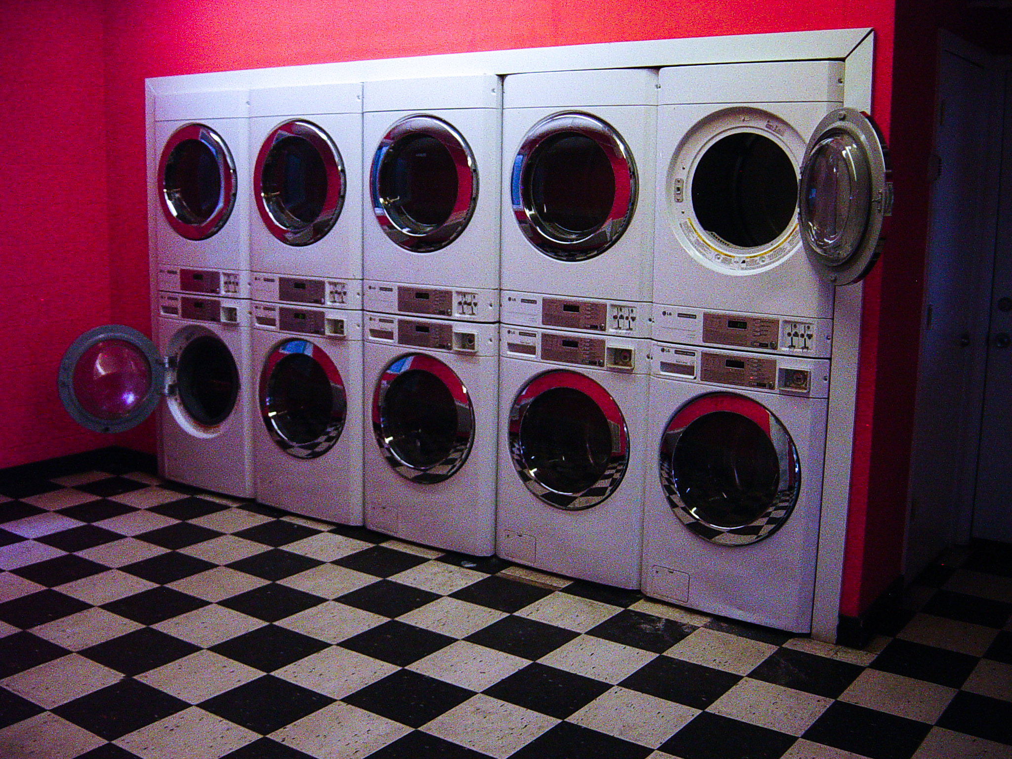 A laundromat wall of round washer doors under pink light, one machine hanging open over black-and-white tile