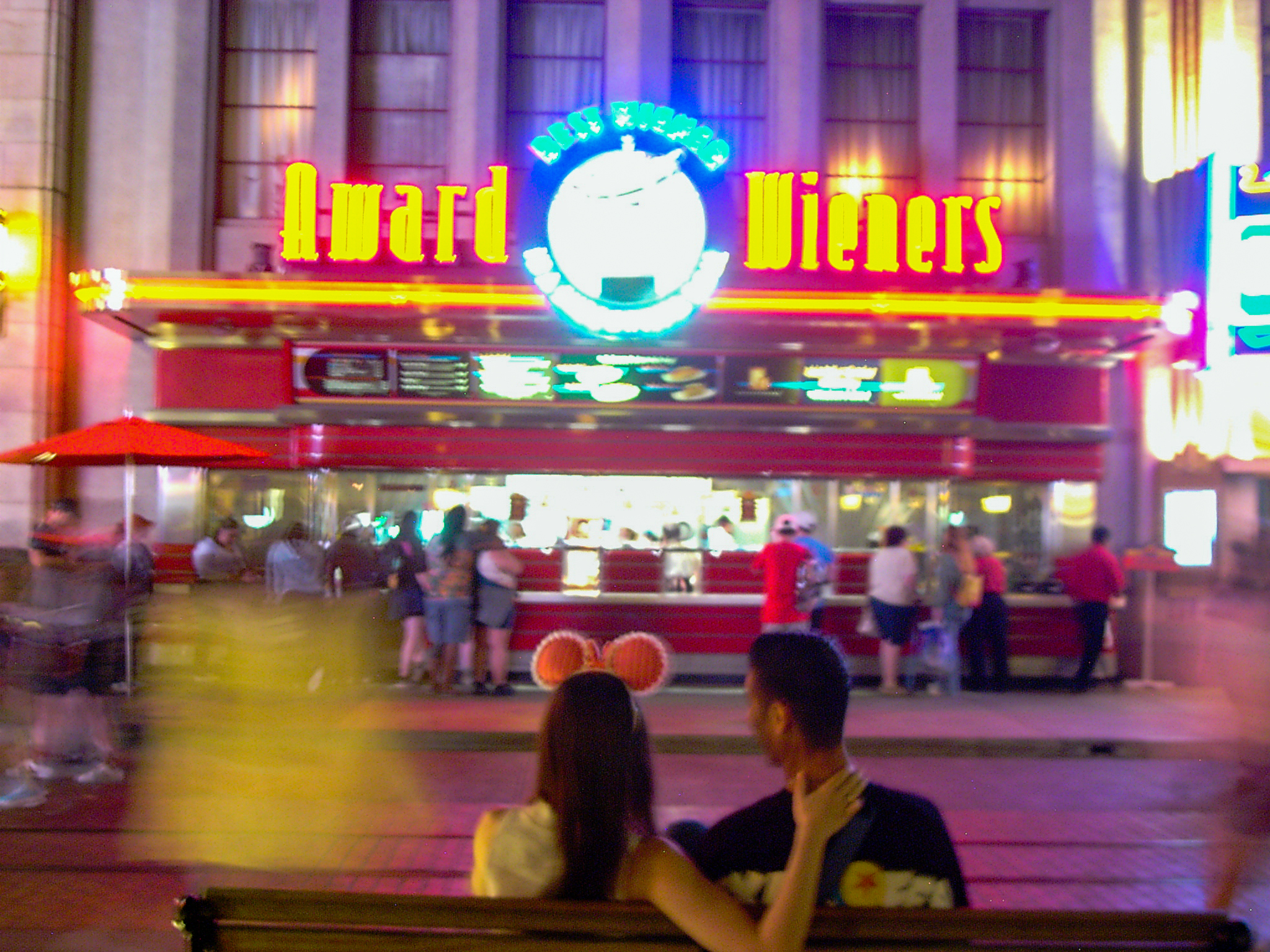 A bright diner sign and passing figures at night, neon smearing into yellow and magenta blur